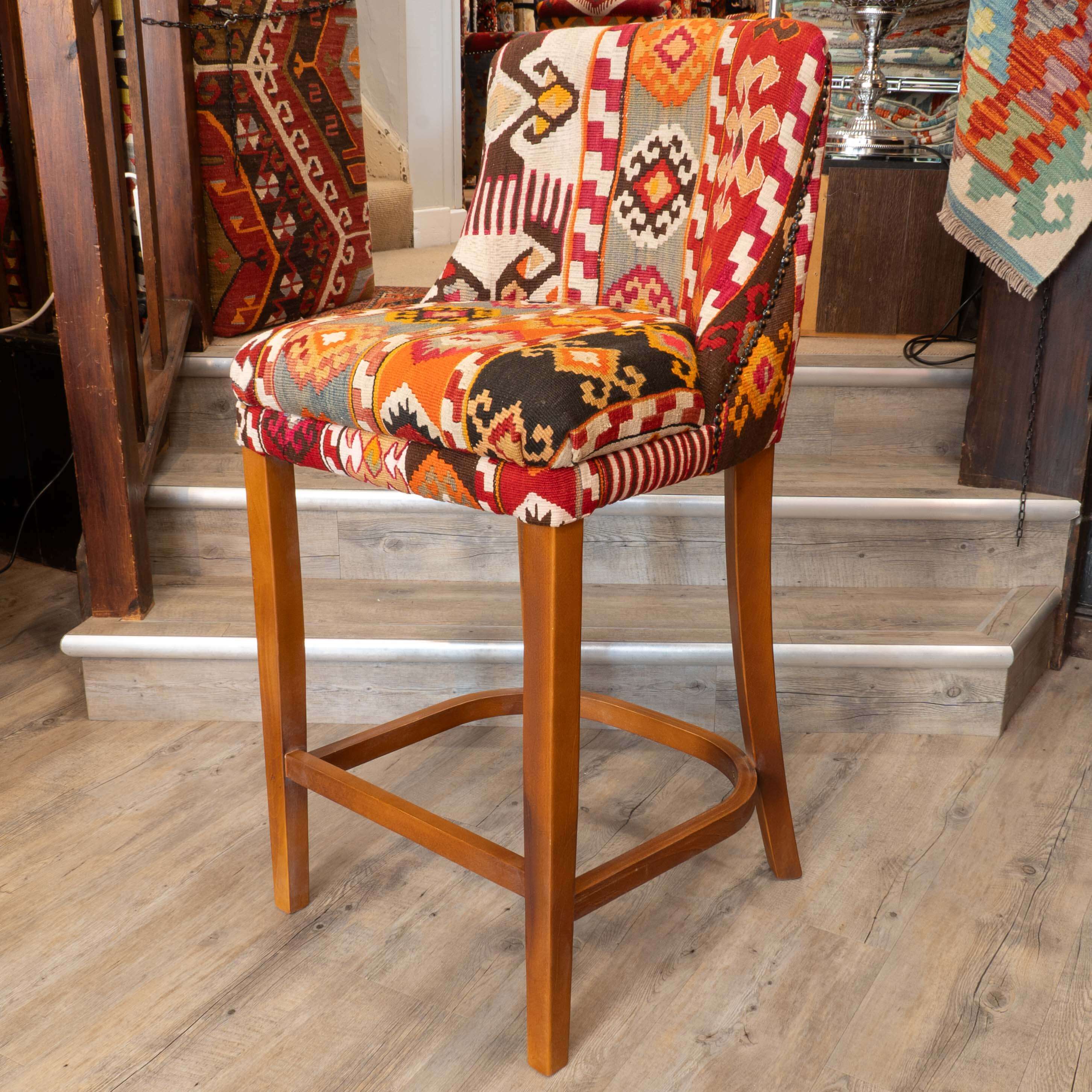 A wooden bar stool with a colorful geometric patchwork fabric upholstery, set in a home interior with wooden stairs and rugs in the background.