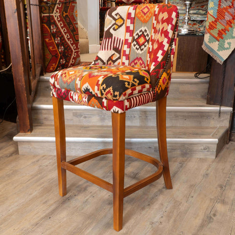 A wooden bar stool with a colorful geometric patchwork fabric upholstery, set in a home interior with wooden stairs and rugs in the background.