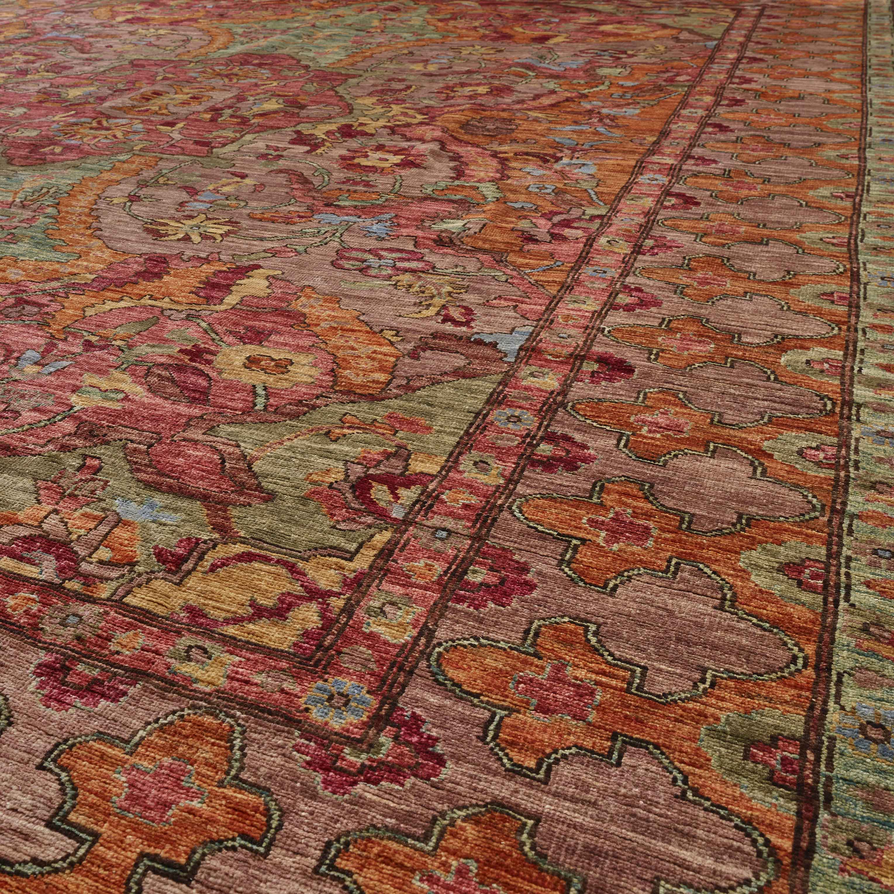 A close-up view of an ornate traditional area rug with a complex floral and geometric pattern in warm tones of rust orange, dusty pink, olive green, and burgundy.