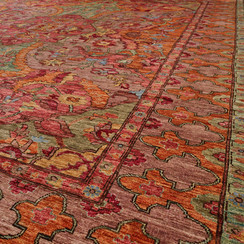 A close-up view of an ornate traditional area rug with a complex floral and geometric pattern in warm tones of rust orange, dusty pink, olive green, and burgundy.