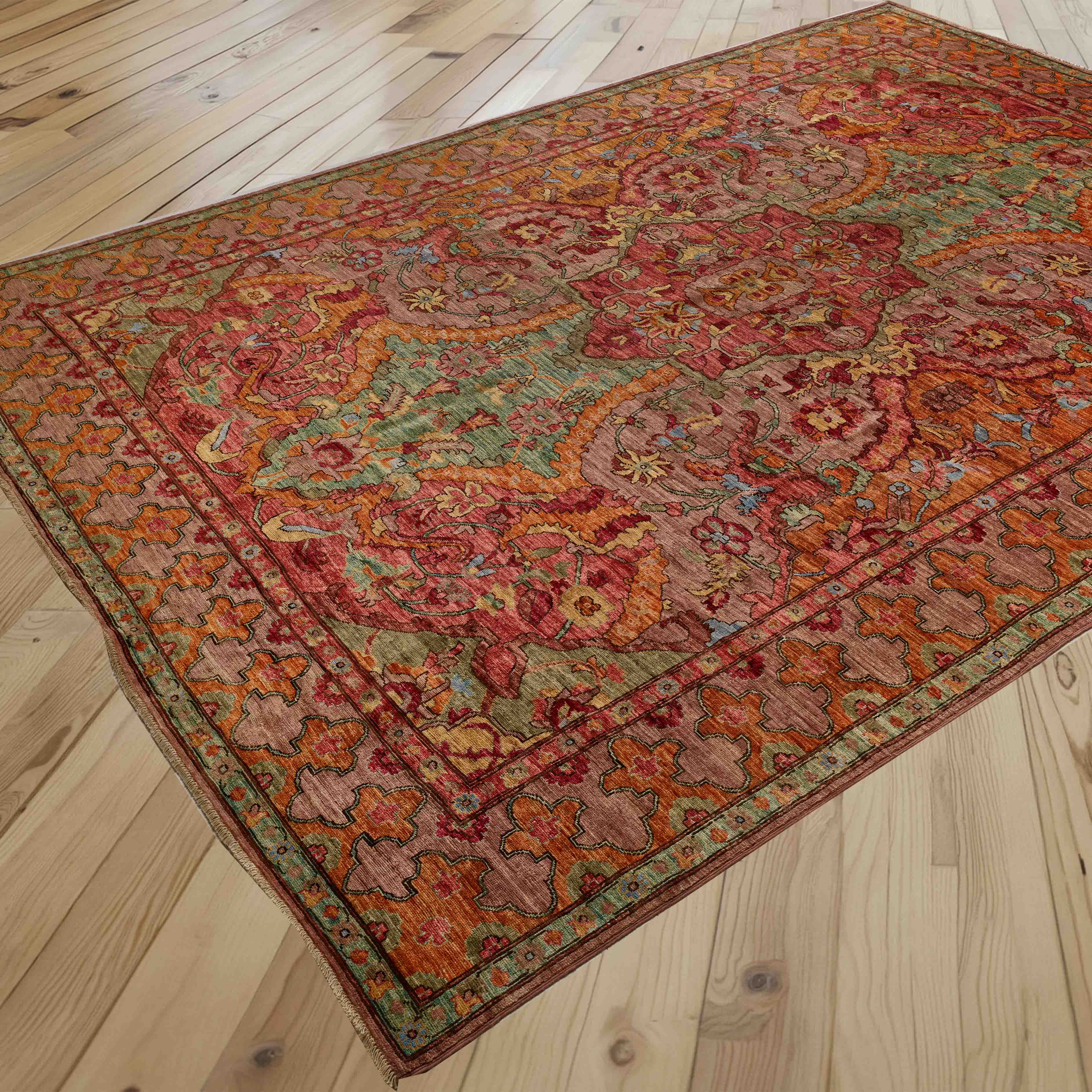 A traditional oriental area rug with an intricate floral and medallion pattern in soft pinkish red, marmalade, soft green, and touches of crushed raspberry, displayed on a light-coloured wooden plank floor.