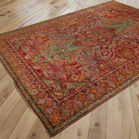 A traditional oriental area rug with an intricate floral and medallion pattern in soft pinkish red, marmalade, soft green, and touches of crushed raspberry, displayed on a light-coloured wooden plank floor.