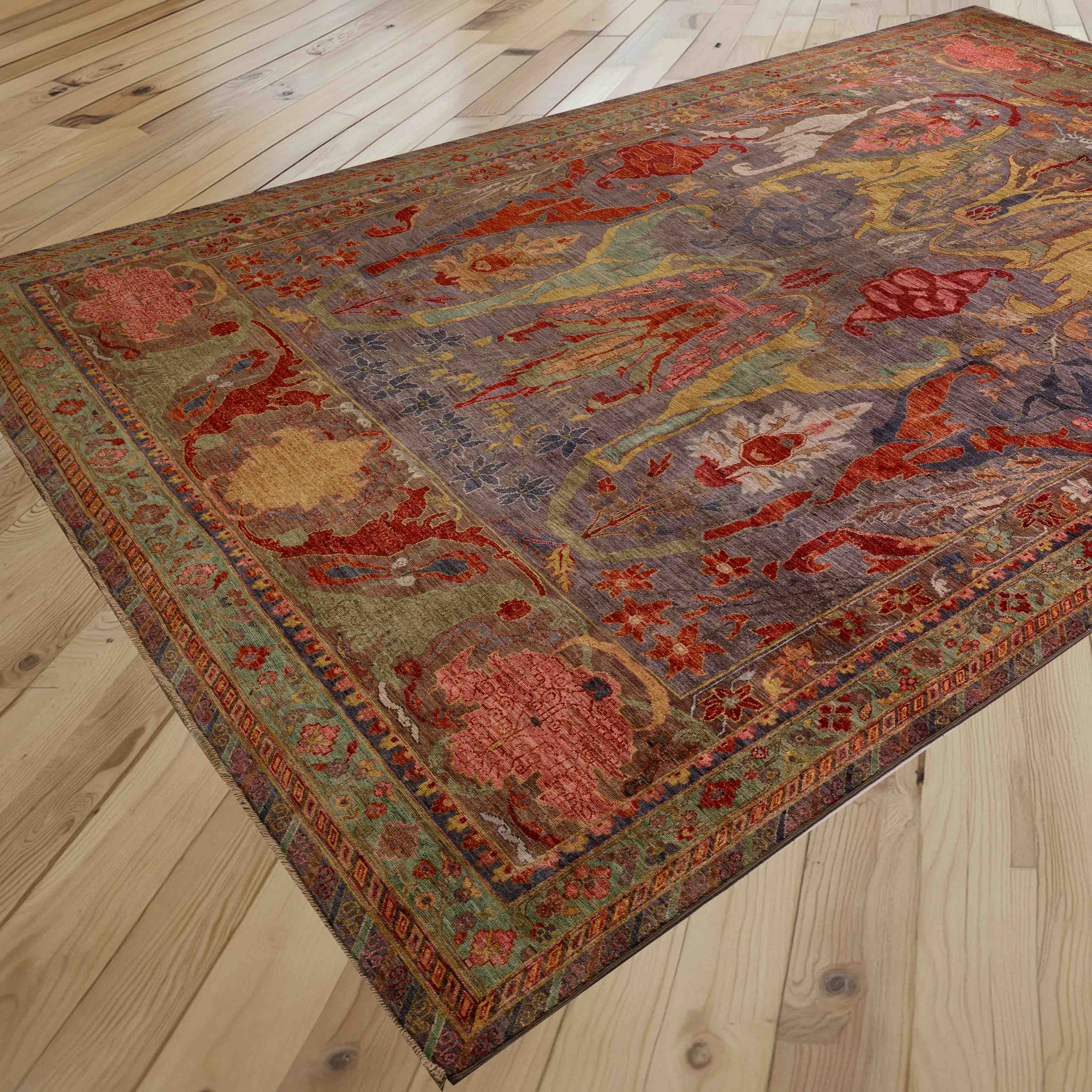 A rectangular area rug with an intricate traditional pattern in burnt orange, grey, soft green, gold and rose pink, shown laid out on a light wood plank floor.