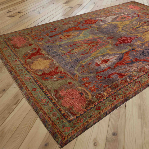 A rectangular area rug with an intricate traditional pattern in burnt orange, grey, soft green, gold and rose pink, shown laid out on a light wood plank floor.