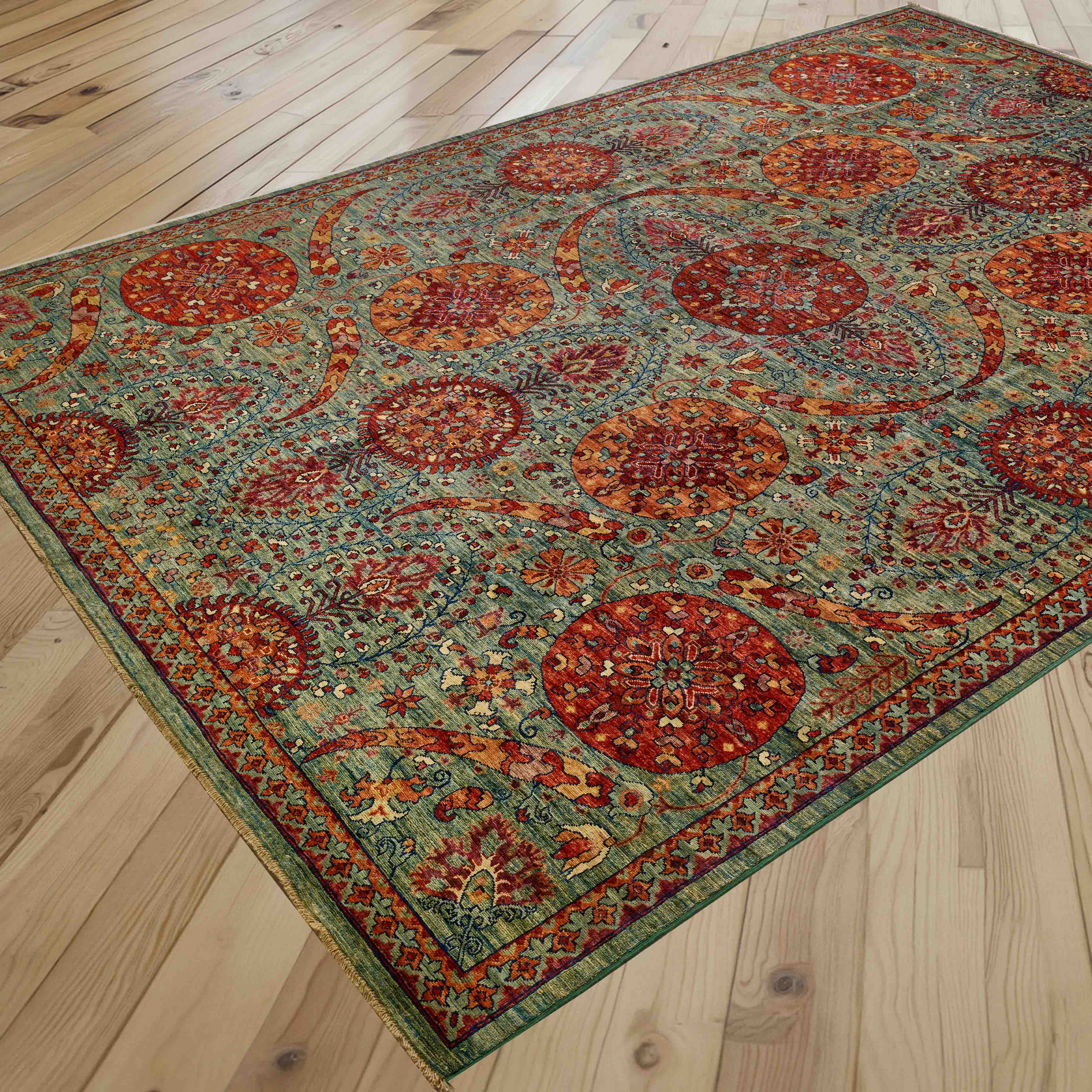 An ornate rectangular area rug with an unusual floral design in green and autumnal colours, shown laid out on a light-coloured wooden plank floor.