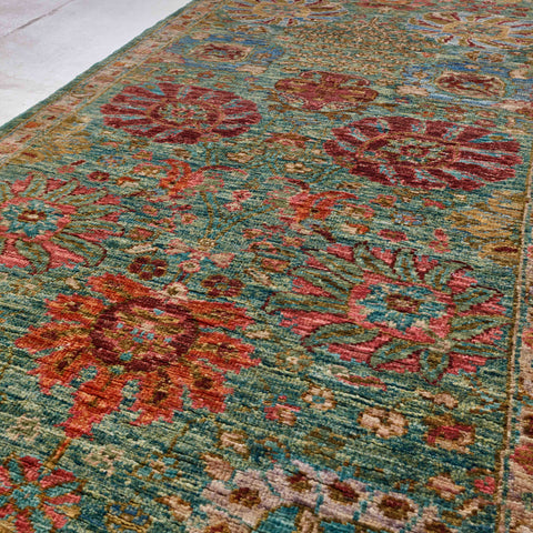 A close-up view of a traditional patterned runner with a teal blue-green base and large floral motifs in papaya spice, hibiscus pink, crimson, and muted gold.