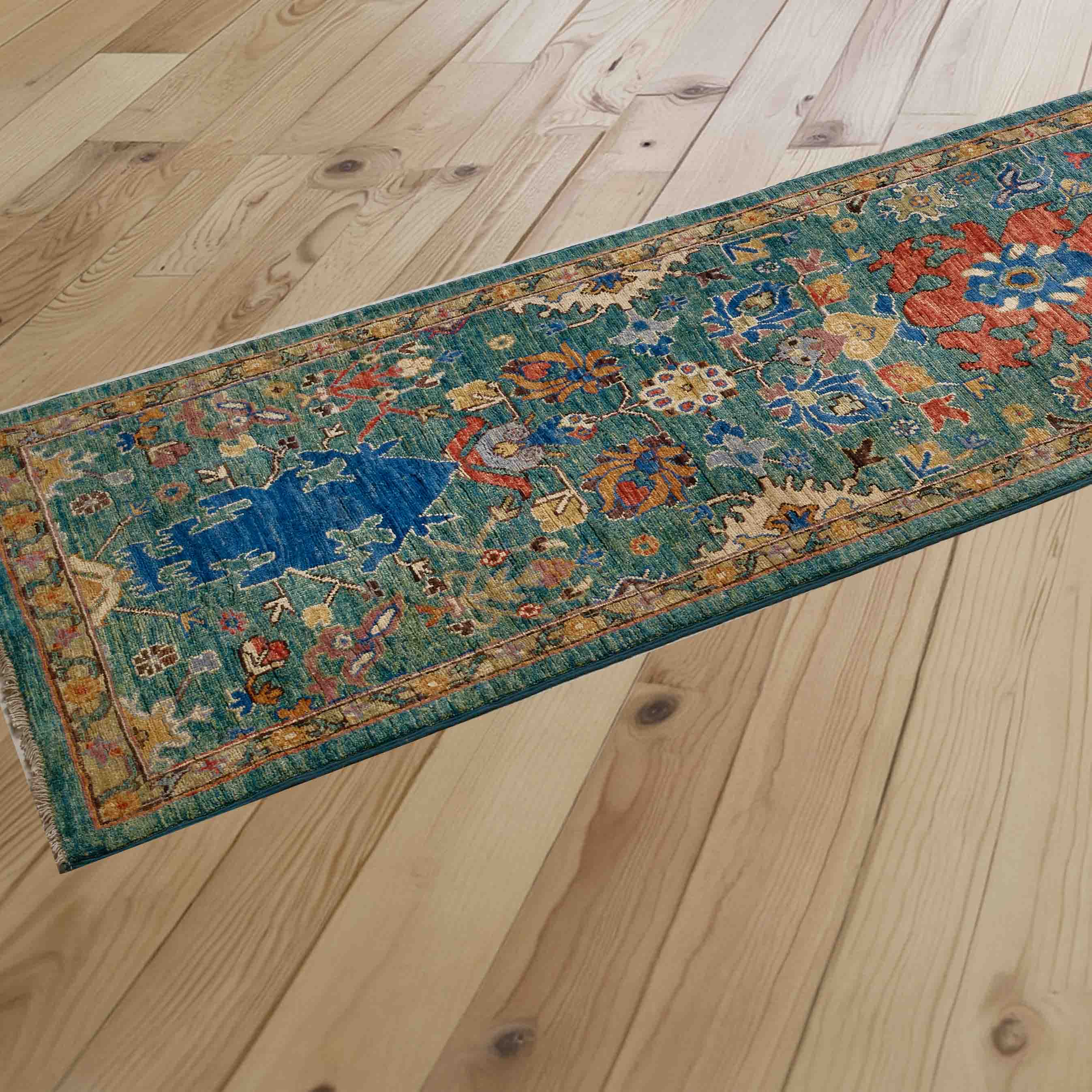 A long runner rug with a distressed traditional pattern in teal, blue, papaya, and honey, laid out diagonally on a light-colored wooden plank floor.