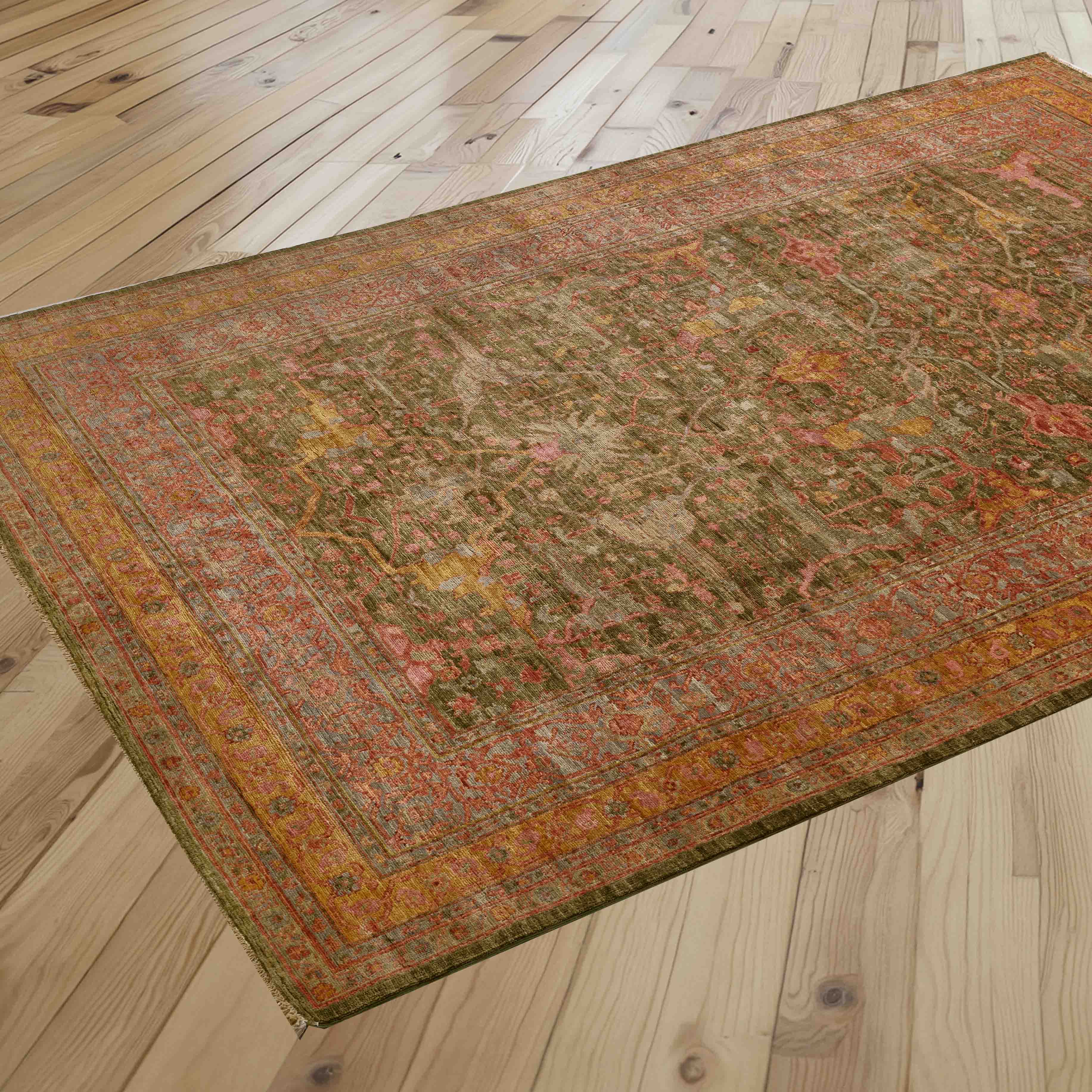 A rectangular vintage-style area rug with an intricate distressed pattern in olive green,  rose pink, soft gold, tangerine and crushed raspberry is shown laid out on a light-colored wooden floor.