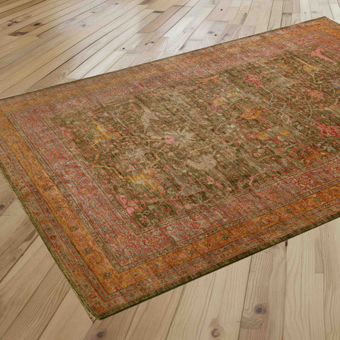 A rectangular vintage-style area rug with an intricate distressed pattern in olive green,  rose pink, soft gold, tangerine and crushed raspberry is shown laid out on a light-colored wooden floor.