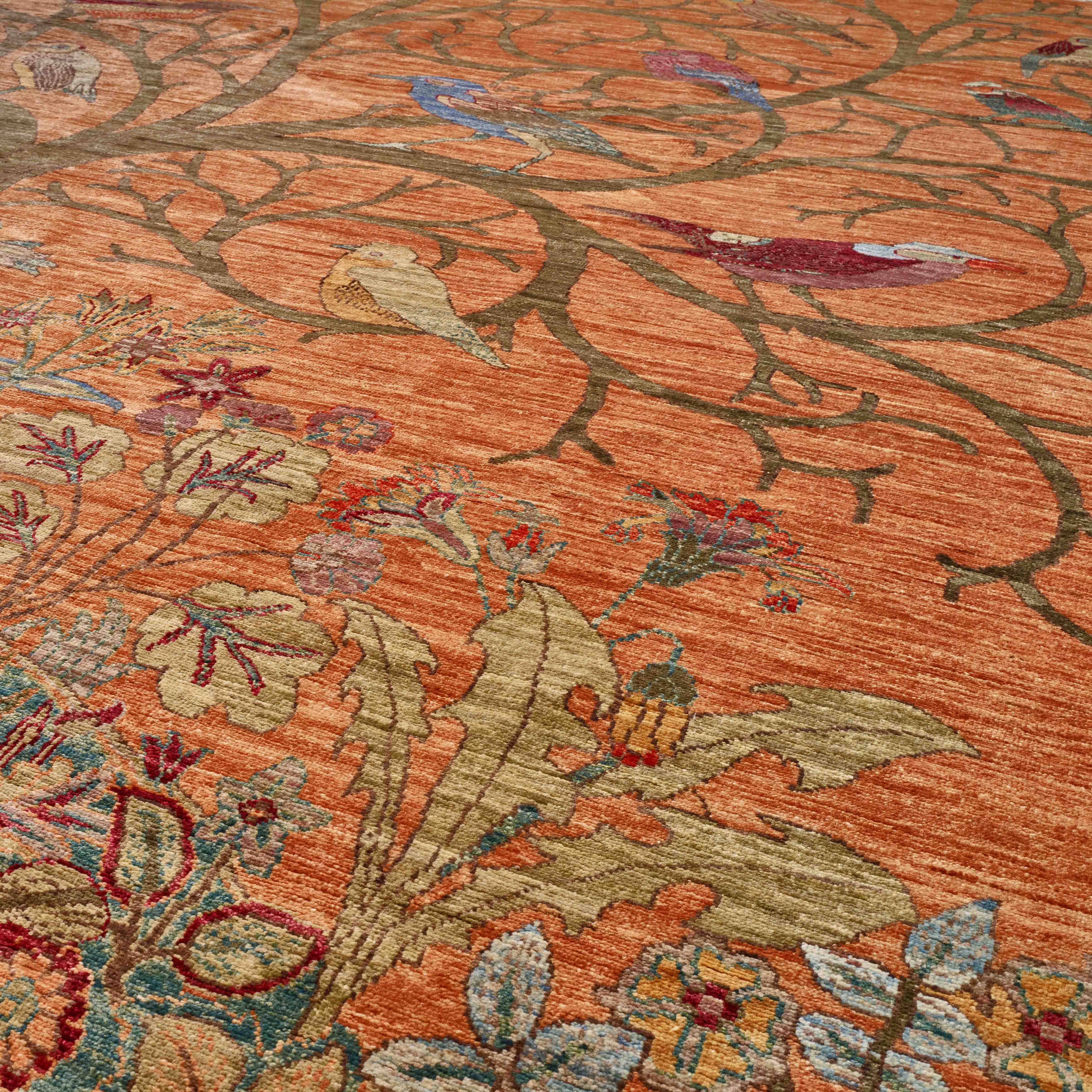 A close-up of an ornate area rug featuring a warm rust-orange background with intricate floral and branch patterns in muted green, beige, red, and blue.