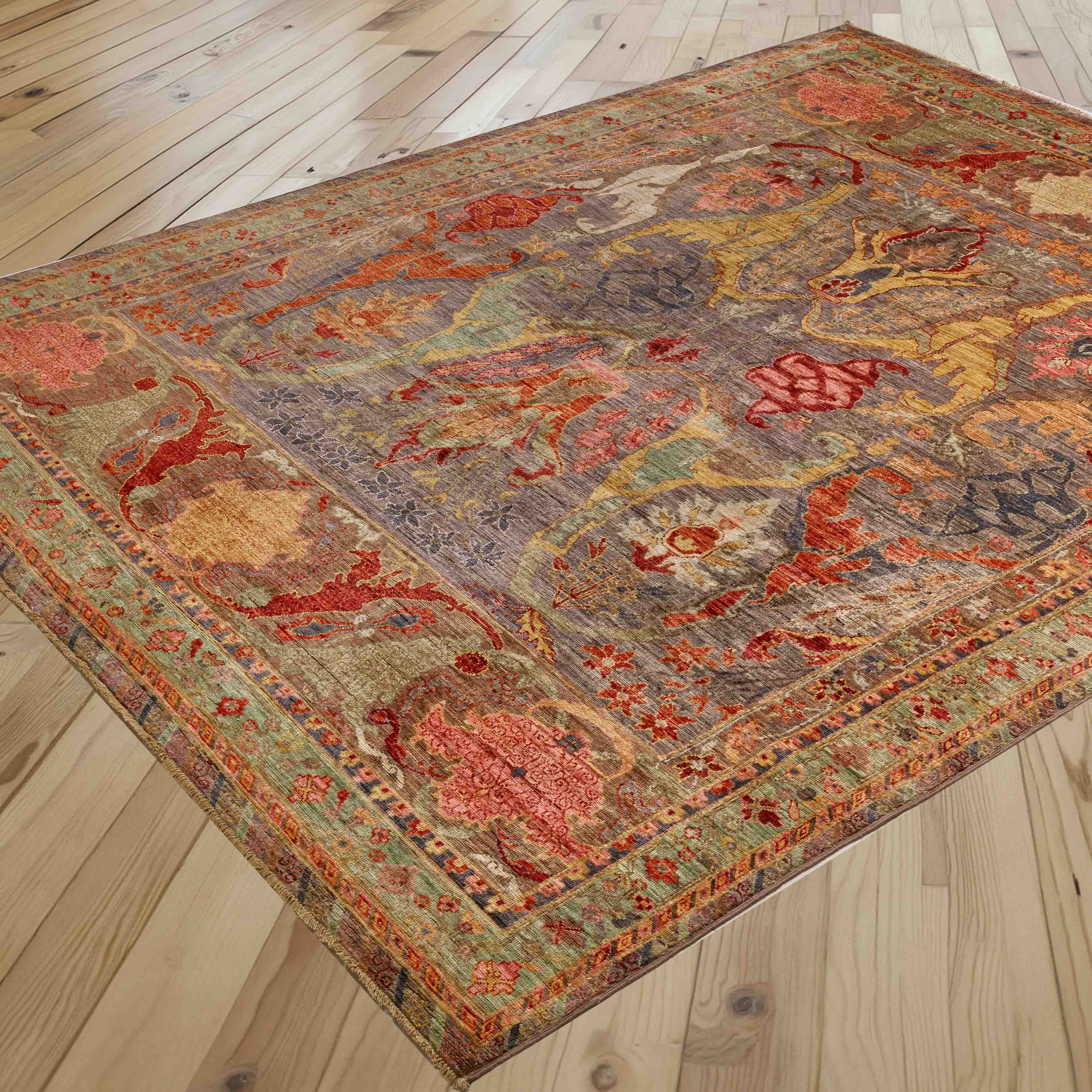 An area rug with an intricate, distressed traditional pattern in muted tones of burnt orange, soft grey, pale green and rose pink is shown laid out on a light wood plank floor.