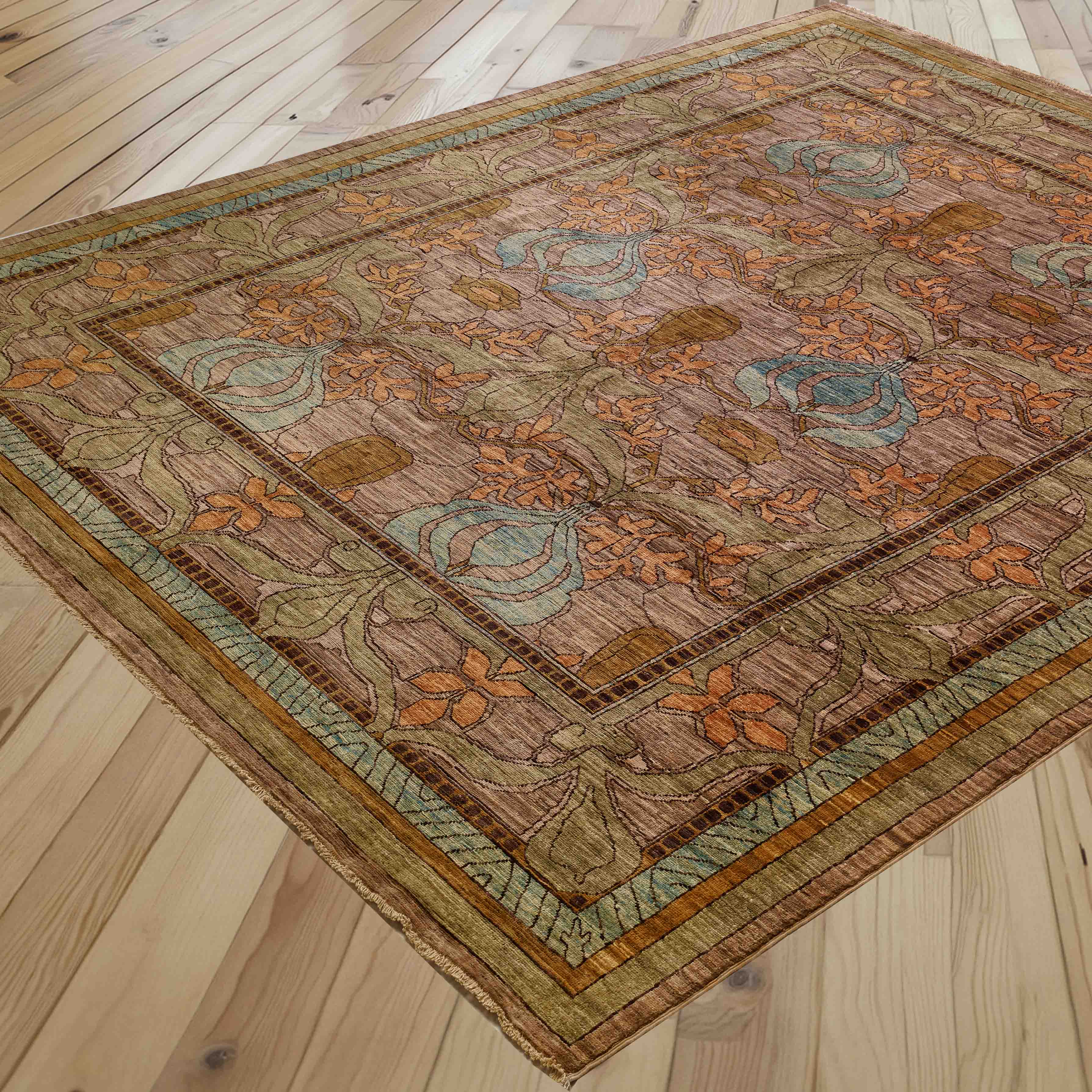 A rectangular area rug with an ornate floral and vine pattern, shown laid out on a light-colored wooden plank floor. The rug features a warm brown base with prominent orange, sage green, and light blue accents.