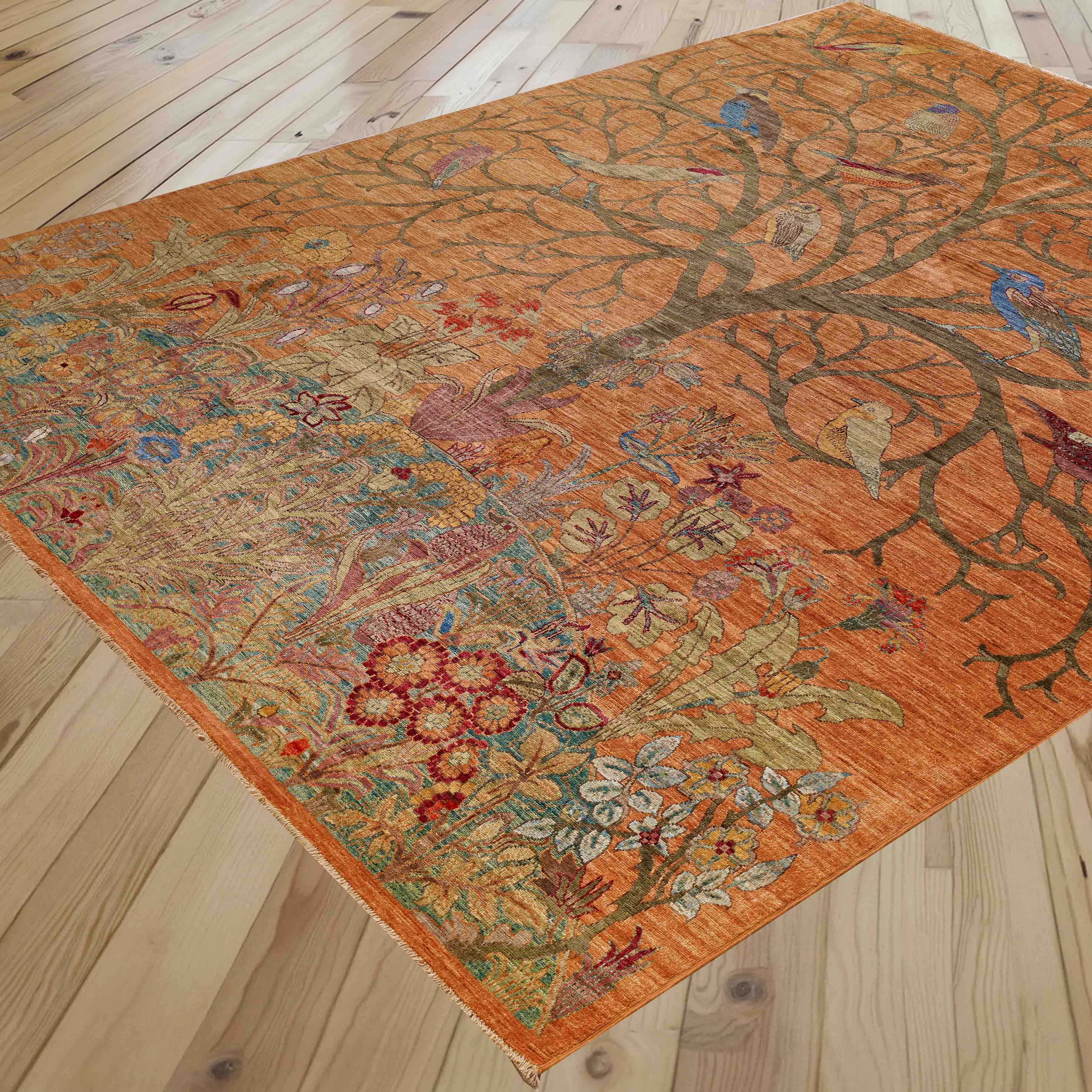 An ornate rectangular area rug with a warm orange-brown base, featuring a detailed botanical and bird pattern, shown laid out on a light wood plank floor.