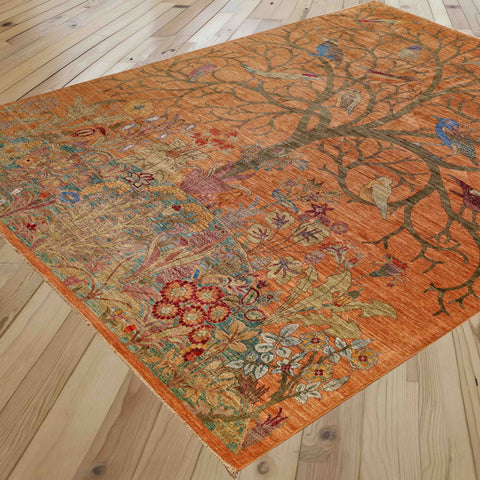 An ornate rectangular area rug with a warm orange-brown base, featuring a detailed botanical and bird pattern, shown laid out on a light wood plank floor.