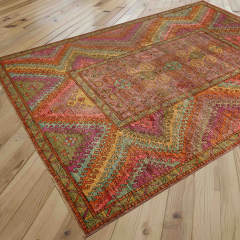 A rectangular area rug with a complex geometric and tribal pattern in multiple warm colors, displayed on a light-colored wooden plank floor.