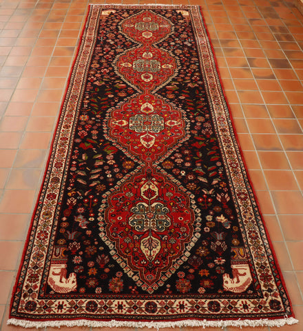 A deep blue ground holds 4 connected leaf shapes in the main area going through the length, these are in broght red.  There is a border in cream. In each corner there is a horses head in cream.  All over, all areas of this rug are depictions of flowers, birds and butterflies. 