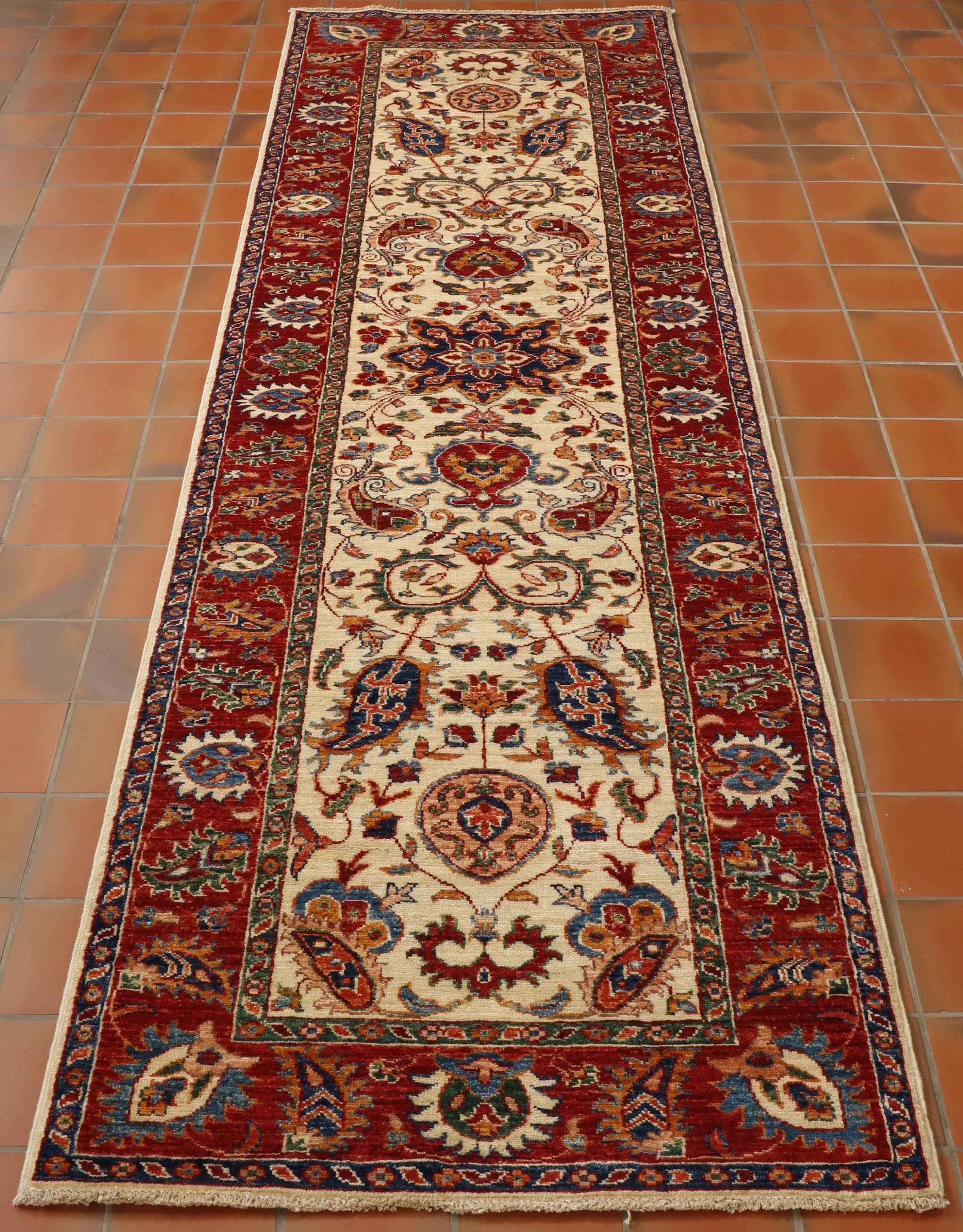 A striking Aryana runner with a cream central ground with flora and fauna design using greens, blues orange, red, peach and golden yellow.  There is a broad red border with contrasting design using colours from the same colour pallet. 