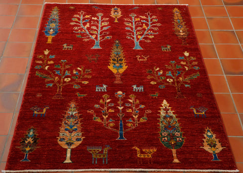 This is a hand knotted rug woven in Afghanistan with a wool pile. The foundation is cotton and it has a short cotton fringe. The background colour is a beautiful rich red and the design is a fairly primitive one with differing styles of trees and random stylised goats and birds. The other colours are old gold, sand, tan, and different shades of blue including turquoise .