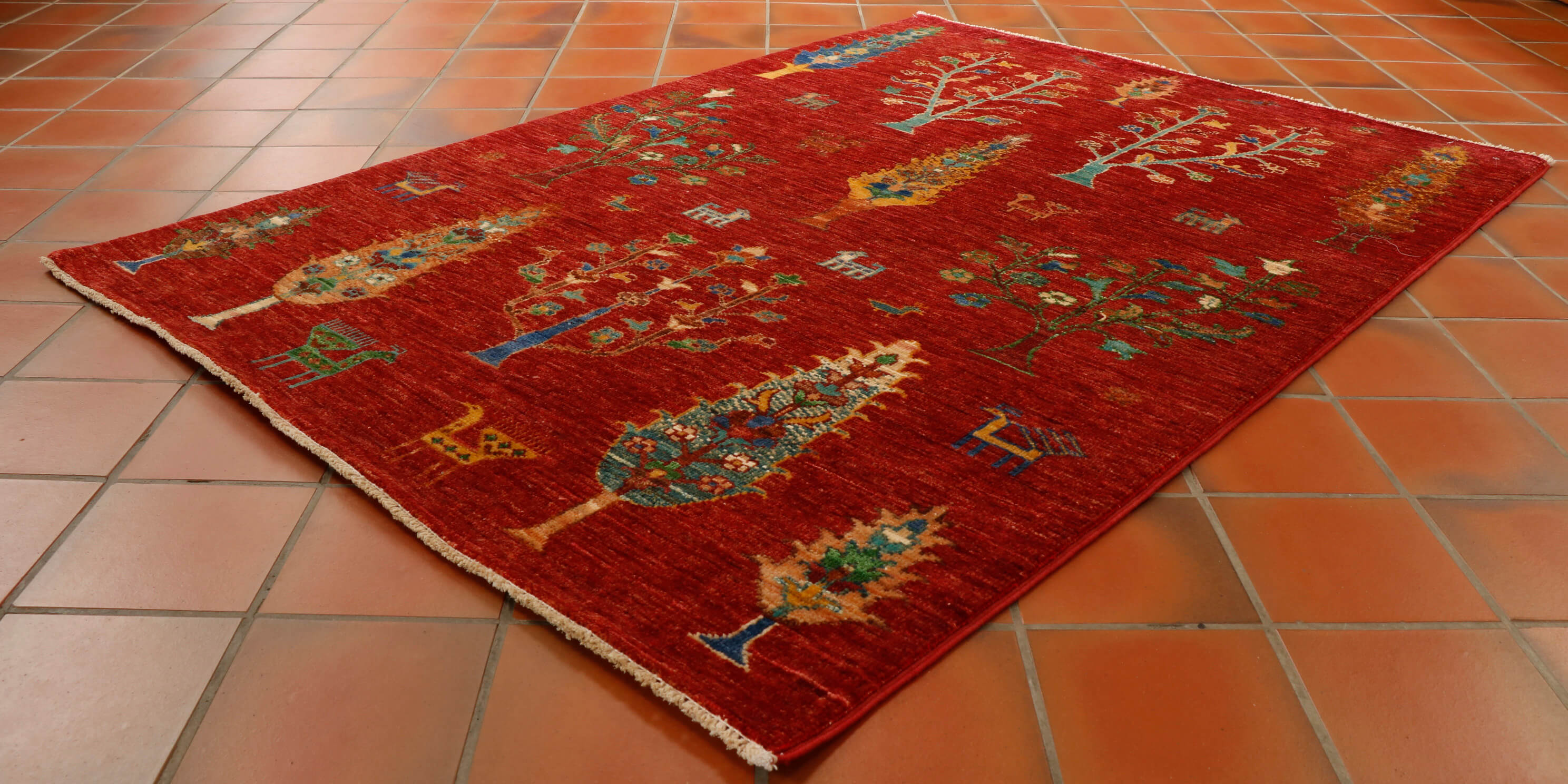 Stunning Afghan Shahi with warm red background and covered in lots of different trees in varying colours of gold, turquoise, blue, green and tan. Random stylised birds and animals have been added to give extra character.