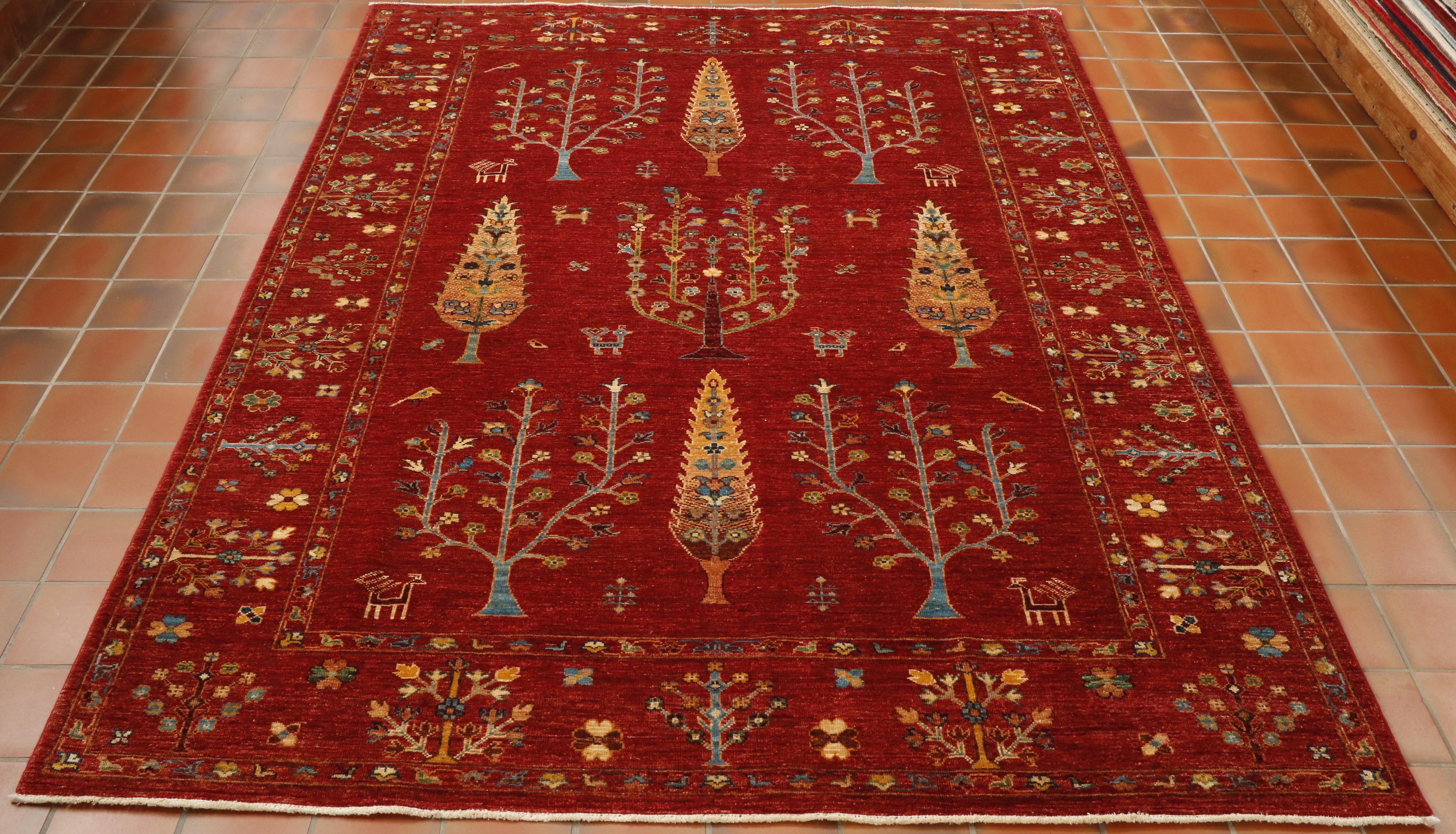 A bright red ground to all of the rug with two types of tree symmetrically laid out upon the central section.  The colours used to show the trees are golden yellow and sea blue.  There are  also some depictions of animals and birds within this section.  In addition to the main colours, greens, browns and oranges are used.  Around the edge of the rug are further tree and floral motifs using the same colour palette, 