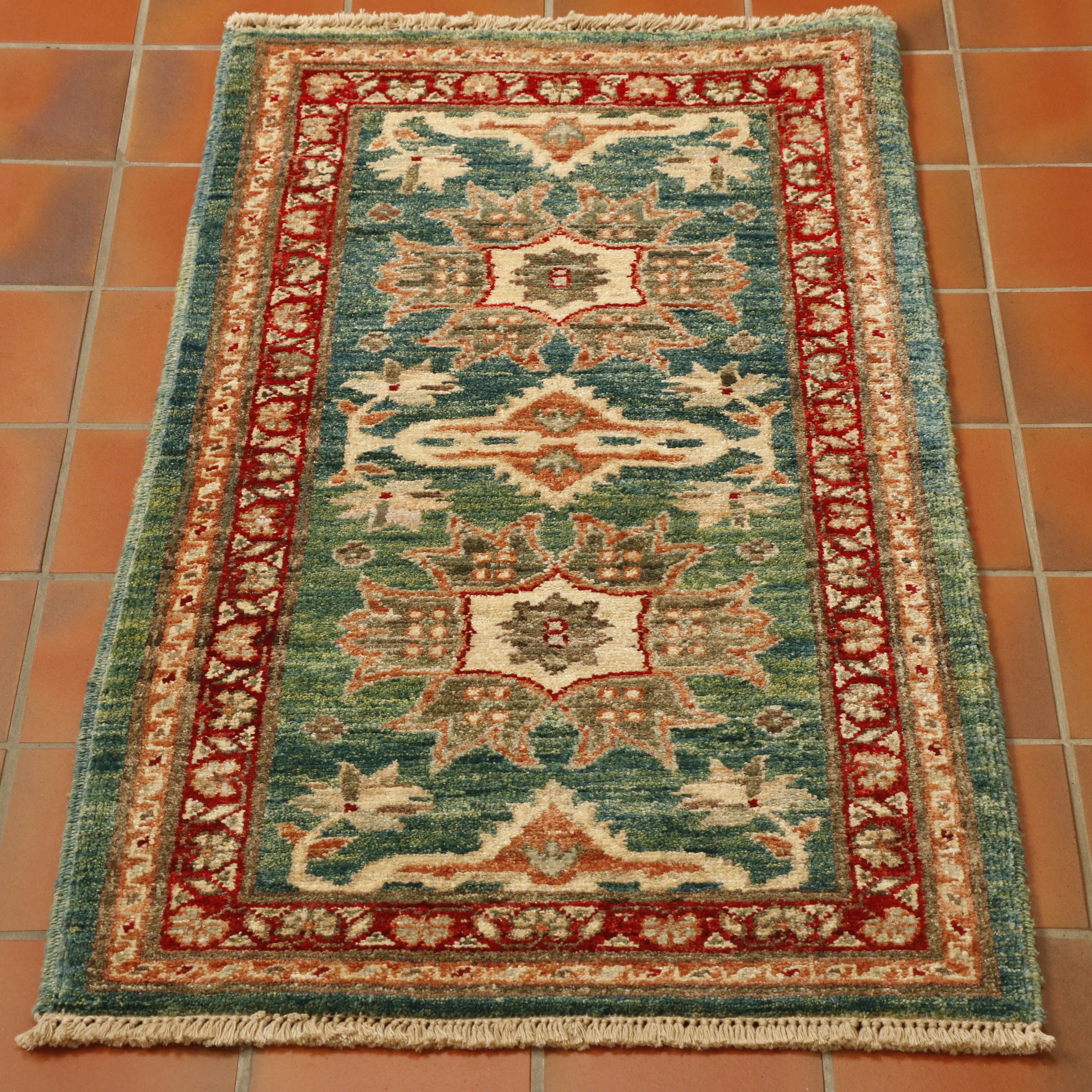 This rug has a greeny blue background, there are parts which are more green than blue. The decoration is stylized flowers.  A main one in the centre, two larger, different ones follow either side and then two half flowers, mirroring the design of the central one at each end.  All of these have a predominantly cream and terracotta palette.  