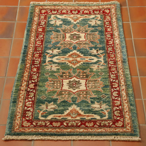 This rug has a greeny blue background, there are parts which are more green than blue. The decoration is stylized flowers.  A main one in the centre, two larger, different ones follow either side and then two half flowers, mirroring the design of the central one at each end.  All of these have a predominantly cream and terracotta palette.  